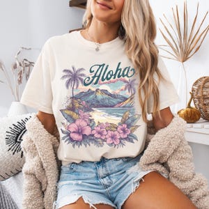 以下が含まれることがあります： クリーム色のTシャツ。トロピカルな風景のデザインで、ターコイズ色のスクリプトで「Aloha」の文字が描かれています。ヤシの木、山、ビーチ、ピンクの花のグラフィック。デニムのショートパンツと合わせています。