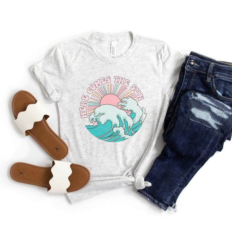 Op de afbeelding: Een grijs t-shirt met een afbeelding van een zon die opkomt boven golven en de tekst "Here Comes The Sun".