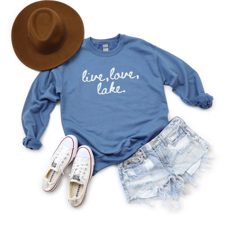 May include: A blue sweatshirt with the text "live, love, lake" in white lettering. The sweatshirt is paired with denim shorts, a brown hat, and white sneakers.