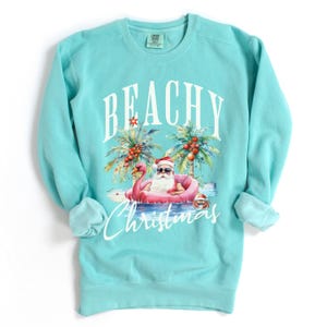May include: A turquoise sweatshirt with the words "BEACHY Christmas" in white script. The graphic features Santa Claus in sunglasses, floating on a pink flamingo in the ocean, with palm trees and red berries. The sweatshirt has a crew neck and long sleeves.