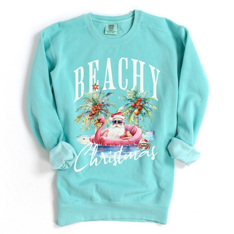 May include: A turquoise sweatshirt with the words "BEACHY Christmas" in white script. The graphic features Santa Claus in sunglasses, floating on a pink flamingo in the ocean, with palm trees and red berries. The sweatshirt has a crew neck and long sleeves.