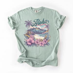 以下が含まれることがあります： トロピカルビーチのシーンが描かれた、薄い緑色のTシャツ。「Aloha」の文字はターコイズ色の筆記体で、ヤシの木、山、ピンクのハイビスカスの花が描かれています。このシャツは柔らかい素材で作られています。