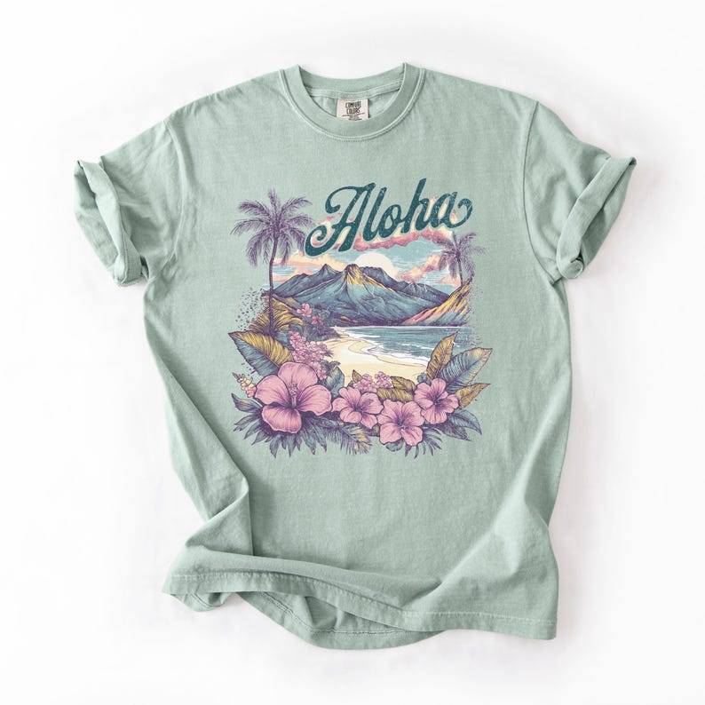 以下が含まれることがあります： トロピカルビーチのシーンが描かれた、薄い緑色のTシャツ。「Aloha」の文字はターコイズ色の筆記体で、ヤシの木、山、ピンクのハイビスカスの花が描かれています。このシャツは柔らかい素材で作られています。