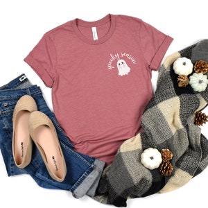 May include: A mauve t-shirt with a white ghost and the text "spooky season" printed on the front. The shirt is laying on a white surface with a pair of jeans, flats, and a plaid blanket.