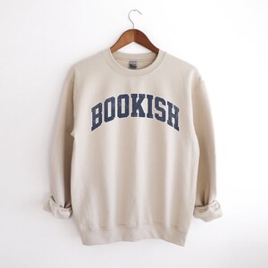 May include: A beige sweatshirt with the word "BOOKISH" printed in a navy blue arch across the front.