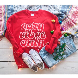 May include: Red sweatshirt with white text that reads "cozy vibes only". The sweatshirt is laid out on a white wooden surface with a pair of white sneakers, blue jeans, and Christmas ornaments.