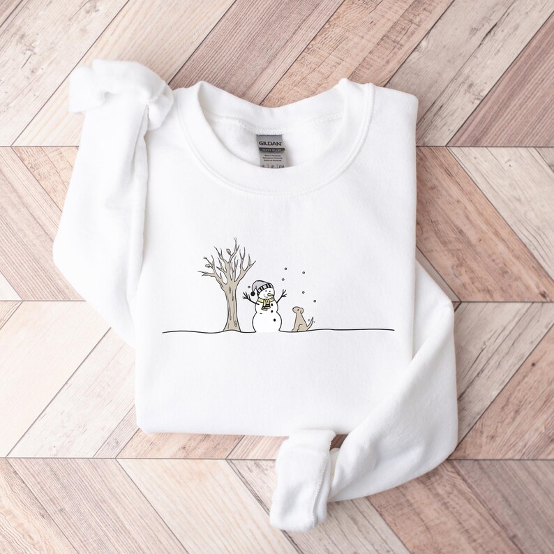 Op de afbeelding: Witte sweatshirt met een winterse illustratie. Een sneeuwman met een hoed en sjaal staat naast een kale boom, met een kleine hond. Het ontwerp is in zwart en bruin, met vallende sneeuw. De sweatshirt is gemaakt van zacht materiaal.
