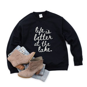 Op de afbeelding: Zwart sweatshirt met de tekst "life is better at the lake." in witte letters. Het sweatshirt ligt plat op een witte achtergrond met een paar bruine enkellaarsjes en een blauwe jeans.