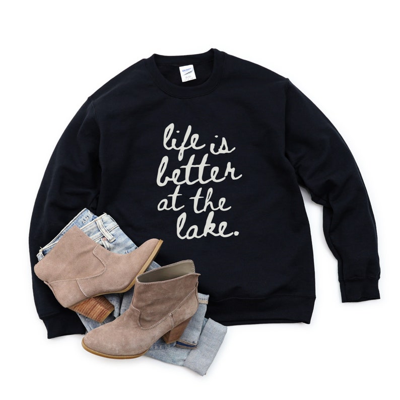 Op de afbeelding: Zwart sweatshirt met de tekst "life is better at the lake." in witte letters. Het sweatshirt ligt plat op een witte achtergrond met een paar bruine enkellaarsjes en een blauwe jeans.