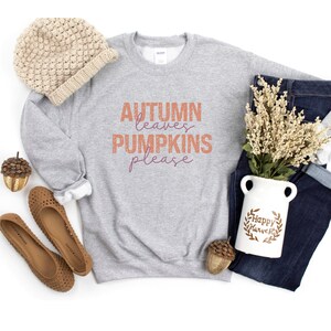 以下が含まれることがあります： オレンジと紫色の「AUTUMN leaves PUMPKINS please」の文字が入ったライトグレーのスウェットシャツ。ベージュのニット帽、茶色のフラットシューズ、「Happy Harvest」の文字が入った白い花瓶、青いジーンズも写っています。