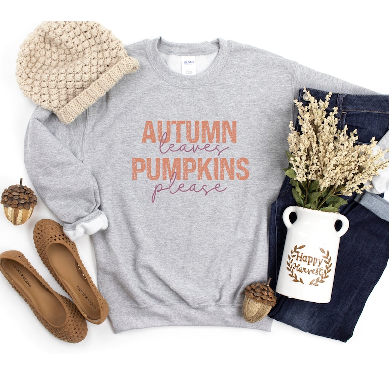 以下が含まれることがあります： オレンジと紫色の「AUTUMN leaves PUMPKINS please」の文字が入ったライトグレーのスウェットシャツ。ベージュのニット帽、茶色のフラットシューズ、「Happy Harvest」の文字が入った白い花瓶、青いジーンズも写っています。