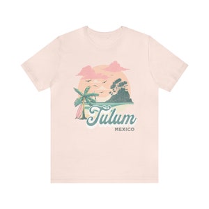 May include: A light pink t-shirt with a graphic design of a sunset over a beach scene with a palm tree and surfboard. The text "Tulum Mexico" is printed below the graphic.