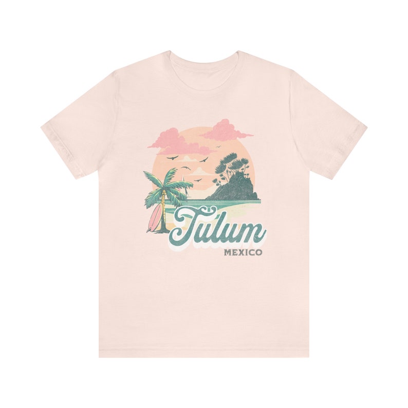 May include: A light pink t-shirt with a graphic design of a sunset over a beach scene with a palm tree and surfboard. The text "Tulum Mexico" is printed below the graphic.