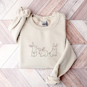 May include: A beige sweatshirt featuring a graphic of three bunnies in a row. The bunnies are white with pink noses and are drawn in a simple, cartoon style.
