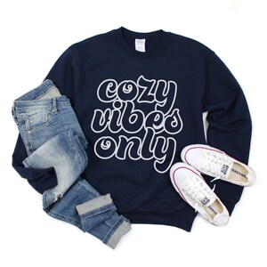 May include: A navy blue sweatshirt with the text "cozy vibes only" in white outlined letters. The sweatshirt is paired with blue jeans and white sneakers.