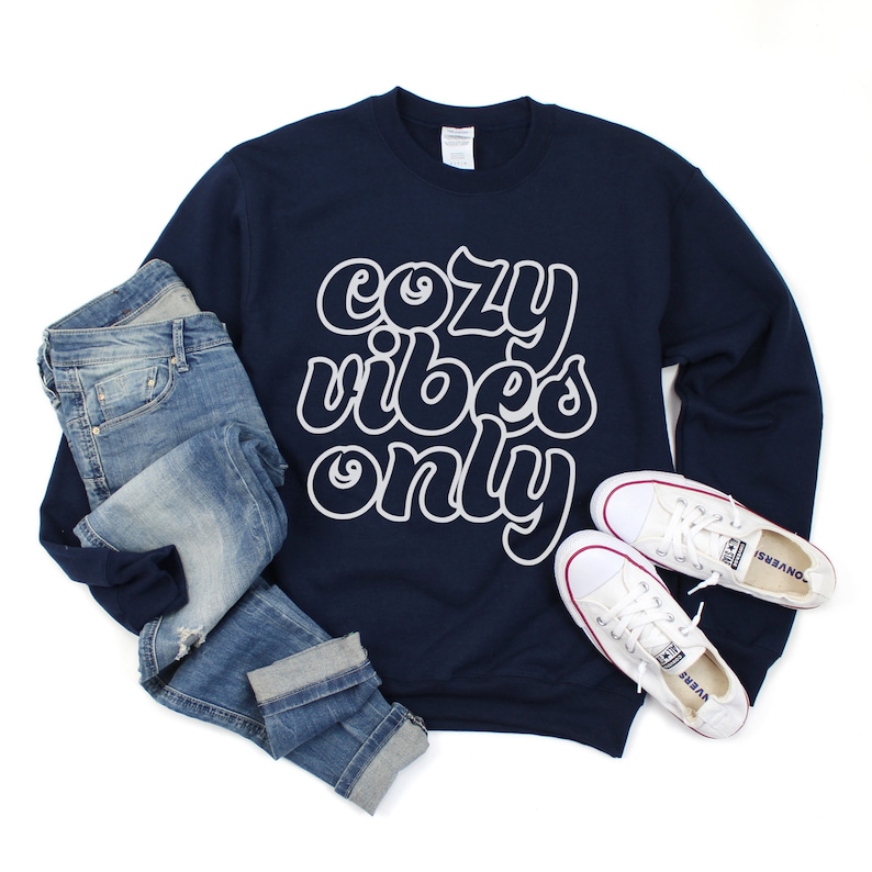 May include: A navy blue sweatshirt with the text "cozy vibes only" in white outlined letters. The sweatshirt is paired with blue jeans and white sneakers.