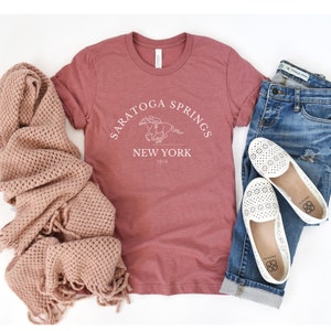 Puede incluir: Una camiseta de color marr&oacute;n rojizo con un dise&ntilde;o gr&aacute;fico blanco que dice "Saratoga Springs New York 1819" y presenta un caballo y un jinete.