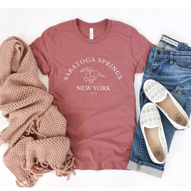 Puede incluir: Una camiseta de color marr&oacute;n rojizo con un dise&ntilde;o gr&aacute;fico blanco que dice "Saratoga Springs New York 1819" y presenta un caballo y un jinete.