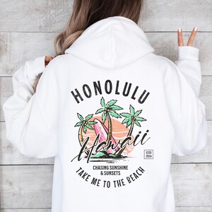 May include: White hoodie with a graphic print of palm trees, a surfboard, and the text "Honolulu Hawaii" in a vintage style. The text "Chasing Sunshine & Sunsets Take Me To The Beach" is printed below the graphic. The text "Estd 2024" is printed in a small font in the bottom right corner.