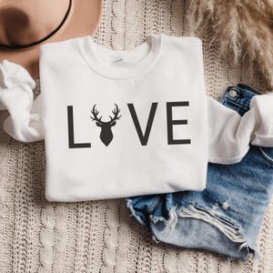 May include: White sweatshirt with the word "LOVE" printed in black letters. The "O" is replaced with a silhouette of a deer head with antlers.