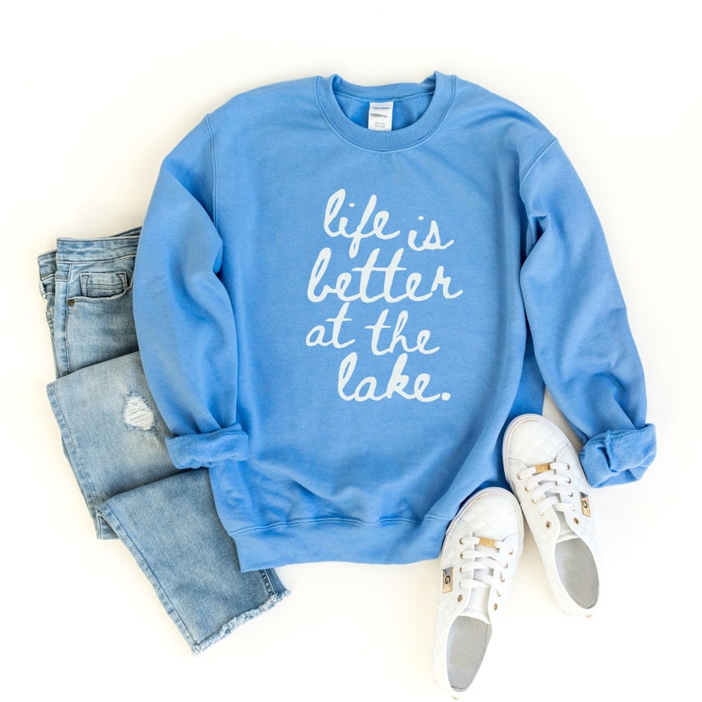 Op de afbeelding: Een lichtblauw sweatshirt met de tekst "life is better at the lake" in witte letters.