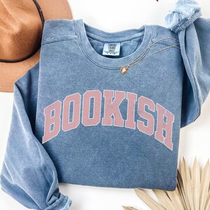 Puede incluir: Una sudadera azul con un gr&aacute;fico rosa "BOOKISH" en una fuente universitaria. La sudadera tiene un aspecto desgastado y vintage.