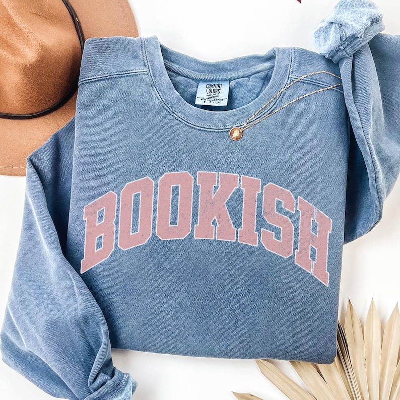 Puede incluir: Una sudadera azul con un gr&aacute;fico rosa "BOOKISH" en una fuente universitaria. La sudadera tiene un aspecto desgastado y vintage.