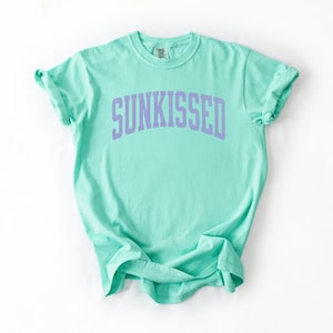 May include: A mint green t-shirt with the word "SUNKISSED" printed in lavender across the chest. The shirt has a crew neck and short sleeves, and is made of a soft, comfortable material.