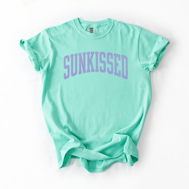 May include: A mint green t-shirt with the word "SUNKISSED" printed in lavender across the chest. The shirt has a crew neck and short sleeves, and is made of a soft, comfortable material.