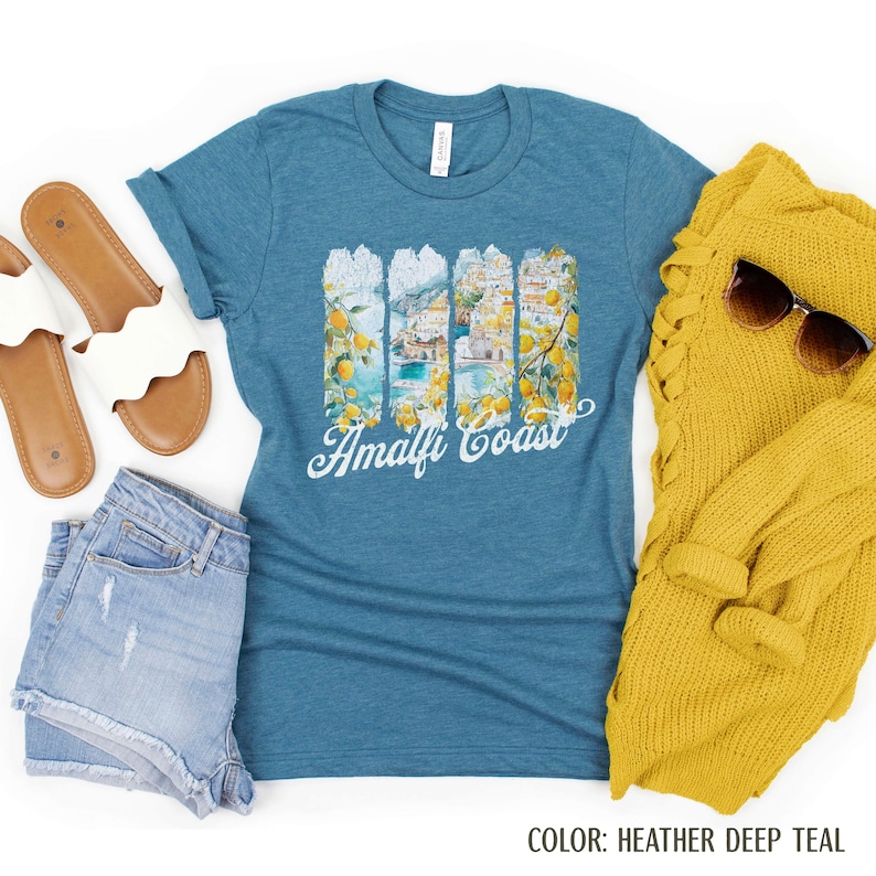 May include: Teal t-shirt with an Amalfi Coast graphic, showcasing lemons and coastal views. The words "Amalfi Coast" are in white script. The shirt is styled with denim shorts, sandals, and a yellow sweater with sunglasses.