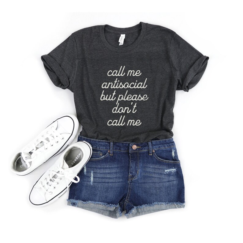 May include: A dark gray t-shirt with the text "call me antisocial but please don't call me" in white lettering. The shirt is paired with denim shorts and white sneakers.