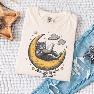 May include: Cream-colored t-shirt featuring a cat reading a book while lounging on a crescent moon. The graphic includes stars, clouds, and the text "Midnight Readers Society." The shirt is paired with blue jeans.