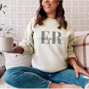 Puede incluir: Sudadera beige claro con el texto "ER nurse" en gris. El dise&ntilde;o incluye una l&iacute;nea de latido card&iacute;aco y un s&iacute;mbolo de coraz&oacute;n. La sudadera se combina con vaqueros azules.