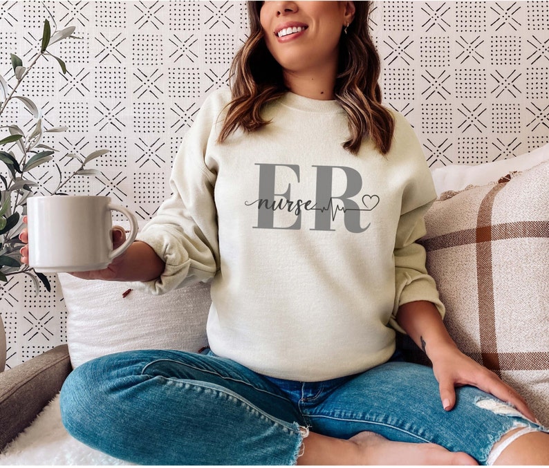 Puede incluir: Sudadera beige claro con el texto "ER nurse" en gris. El dise&ntilde;o incluye una l&iacute;nea de latido card&iacute;aco y un s&iacute;mbolo de coraz&oacute;n. La sudadera se combina con vaqueros azules.