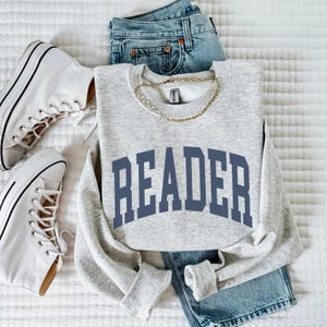 Op de afbeelding: Een grijs sweatshirt met het woord "READER" bedrukt in donkerblauwe letters. Het sweatshirt ligt op een wit oppervlak met een paar witte sneakers en een blauwe jeans.