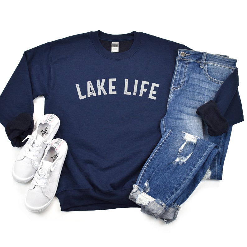 May include: A navy blue sweatshirt with the text "LAKE LIFE" in white lettering. The sweatshirt is paired with blue jeans and white sneakers.