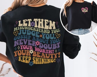 Op de afbeelding: Zwart sweatshirt met de tekst "LET THEM" en een langere inspirerende boodschap in een kleurrijk retro lettertype. Het ontwerp bevat bloemen en hart accenten. Een kleinere versie van het ontwerp staat op de borst.