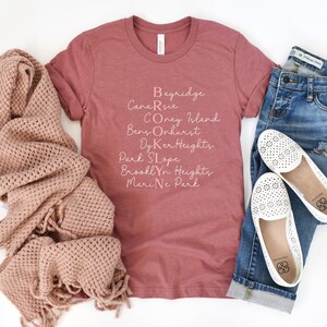 May include: A maroon t-shirt with a pink graphic that says "BROOKLYN" with the names of different neighborhoods in Brooklyn spelled out vertically. The shirt is laid flat on a white surface with a pair of white shoes and a pair of blue jeans.