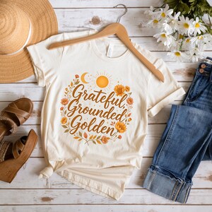 Pu&ograve; includere: T-shirt color crema con le parole "Grateful, Grounded, Golden" in un design floreale circolare. Il design include un sole, una luna e fiori nei toni dell'arancione e del giallo. La maglietta &egrave; esposta su una gruccia di legno.
