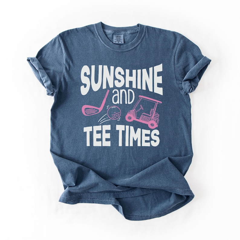 以下が含まれることがあります： 「SUNSHINE and TEE TIMES」の白い文字が入った紺色のTシャツ。ゴルフのクラブ、ゴルフボール、ゴルフカートのピンク色のグラフィックも特徴です。柔らかい素材で作られたシャツです。