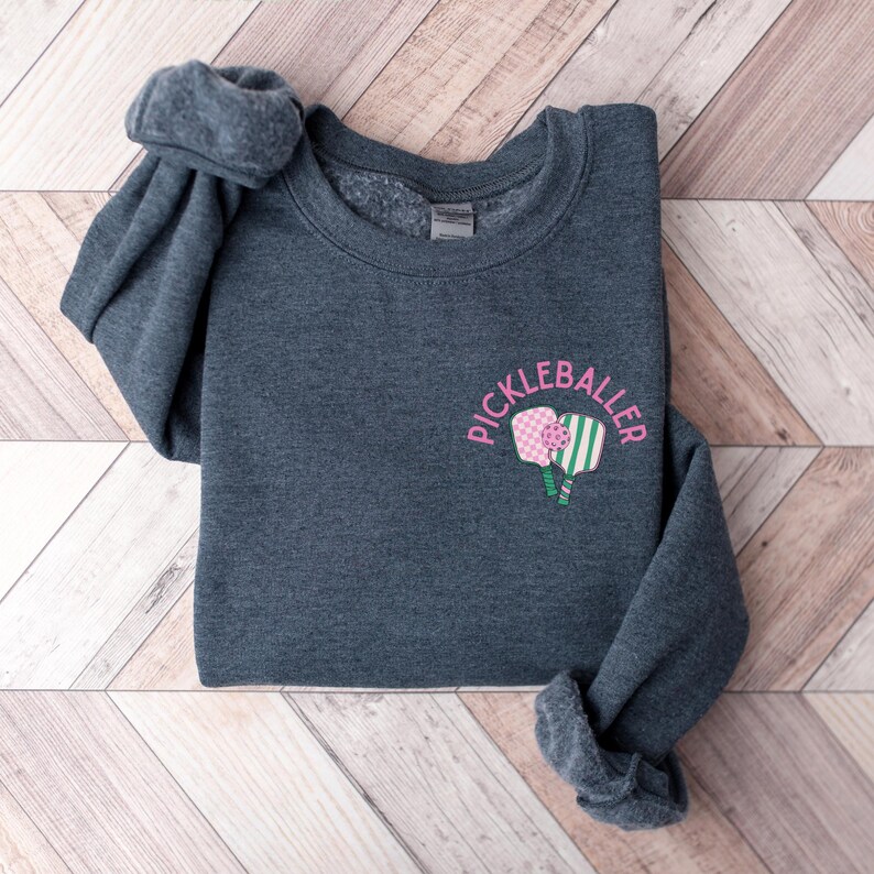 May include: A dark grey sweatshirt with the text "Pickleballer" in pink lettering and a graphic of two pink and green pickleball paddles.