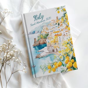 May include: A white travel journal with a watercolor illustration of a coastal Italian town with lemon trees. The text on the cover reads "Italy Travel Adventures 2025".