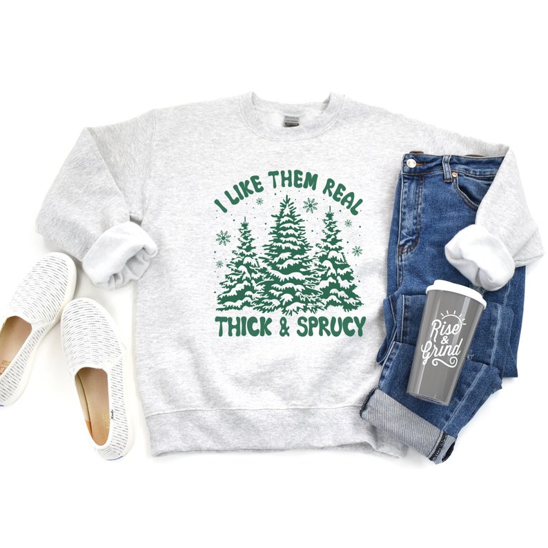 May include: A gray sweatshirt with the text "I like them real thick & sprucy" and a graphic of three green Christmas trees with snow on them. The text is in green and there are white snowflakes around the trees.