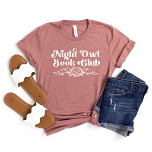 May include: A mauve t-shirt with the text "Night Owl Book Club" in white with a stylized owl graphic. The shirt is laid flat on a white background with a pair of white sandals and denim shorts.