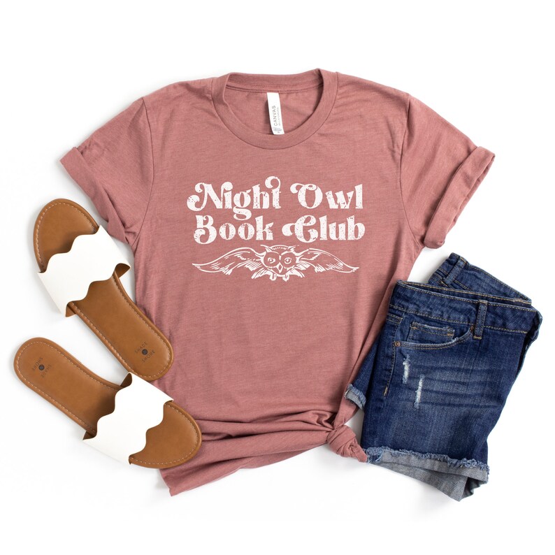 May include: A mauve t-shirt with the text "Night Owl Book Club" in white with a stylized owl graphic. The shirt is laid flat on a white background with a pair of white sandals and denim shorts.