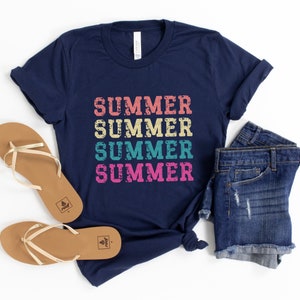 May include: Navy blue t-shirt with the word "SUMMER" printed four times in a retro style, using various colors. The shirt is paired with denim shorts and brown sandals with gold straps, perfect for a summer outfit.