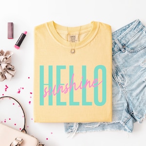 Pu&ograve; includere: T-shirt giallo chiaro con la scritta "HELLO sunshine" in turchese e rosa. La maglietta &egrave; abbinata a pantaloncini di jeans, una collana e trucco. La maglietta &egrave; di Comfort Colors.