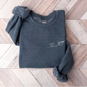 Op de afbeelding: Een donkergrijs sweatshirt met een wit papieren vliegtuigje en een hartvormig ontwerp geborduurd op de voorkant.