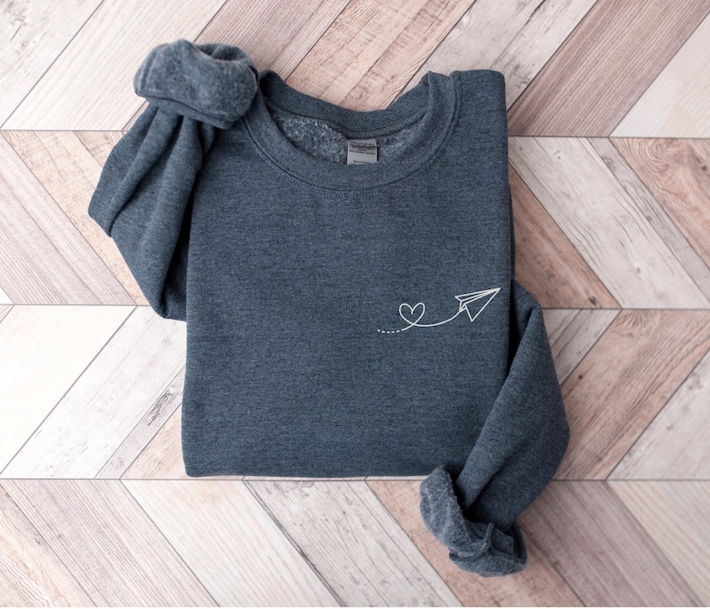 Op de afbeelding: Een donkergrijs sweatshirt met een wit papieren vliegtuigje en een hartvormig ontwerp geborduurd op de voorkant.