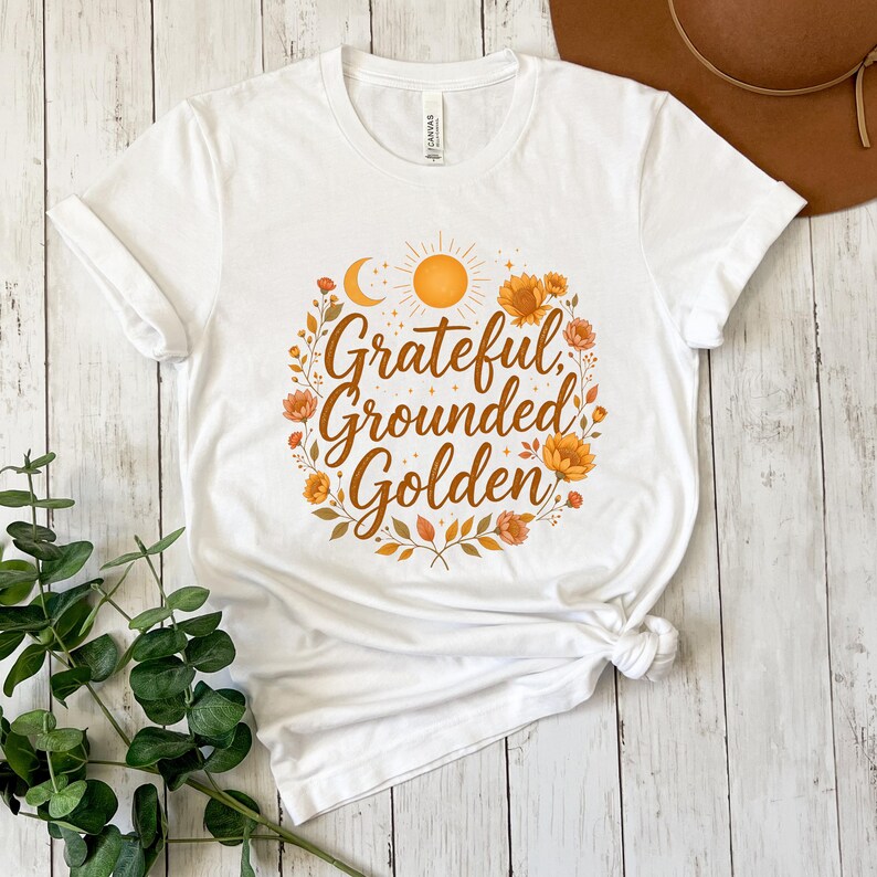Pu&ograve; includere: T-shirt bianca con le parole "Grateful, Grounded, Golden" in un design circolare. Il design include un sole, una luna ed elementi floreali nei toni dell'arancione e del giallo. La maglietta &egrave; realizzata in un materiale morbido.